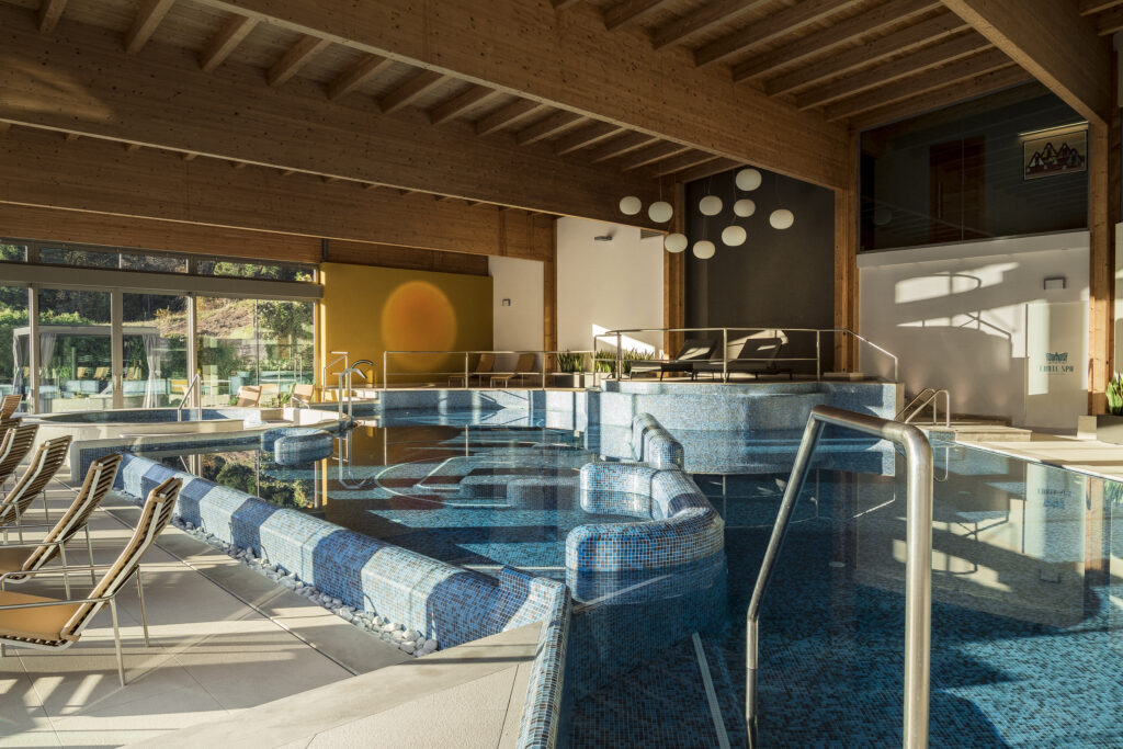 Indoor pool at Day Spa Cadore Dolomiti, featuring blue mosaic tiles and wooden beam ceiling.